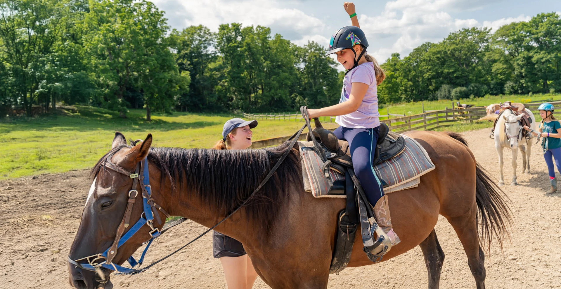 A young girl wearing a helmet rides a horse in a sunny outdoor equestrian area with green trees in the background.