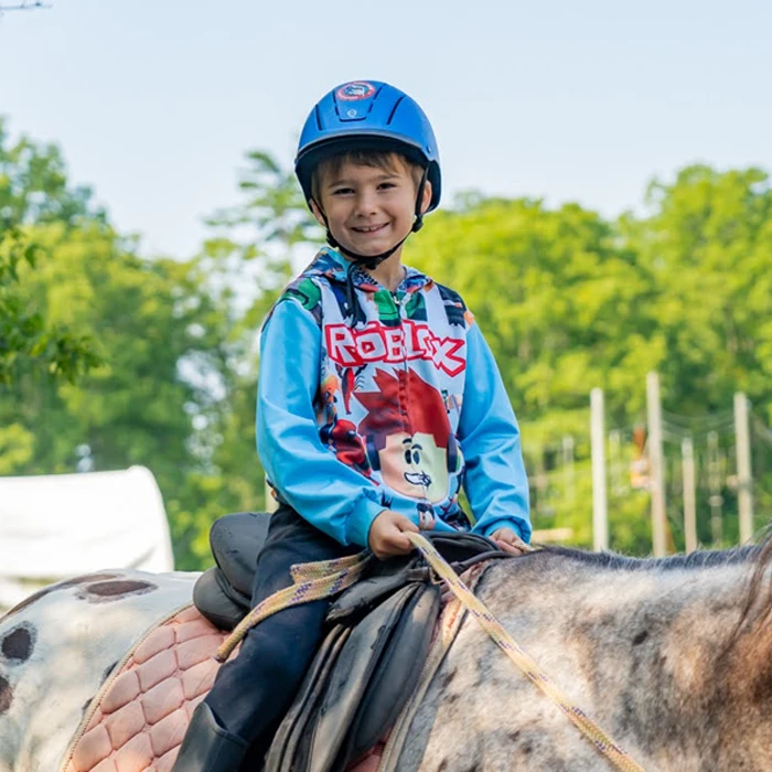 A young boy wearing a helmet rides a horse in a sunny outdoor equestrian area with green trees in the background.