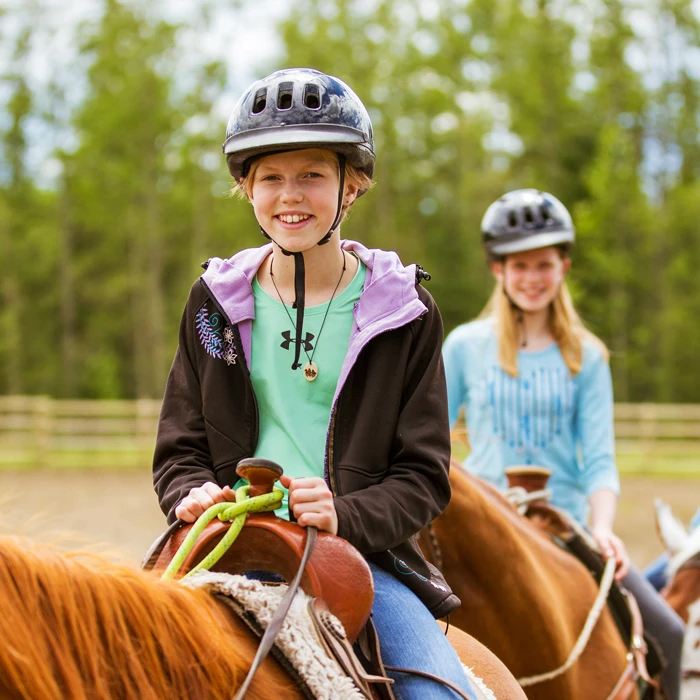 Young girls wearing helmets ride horses in a sunny outdoor equestrian area with green trees in the background.