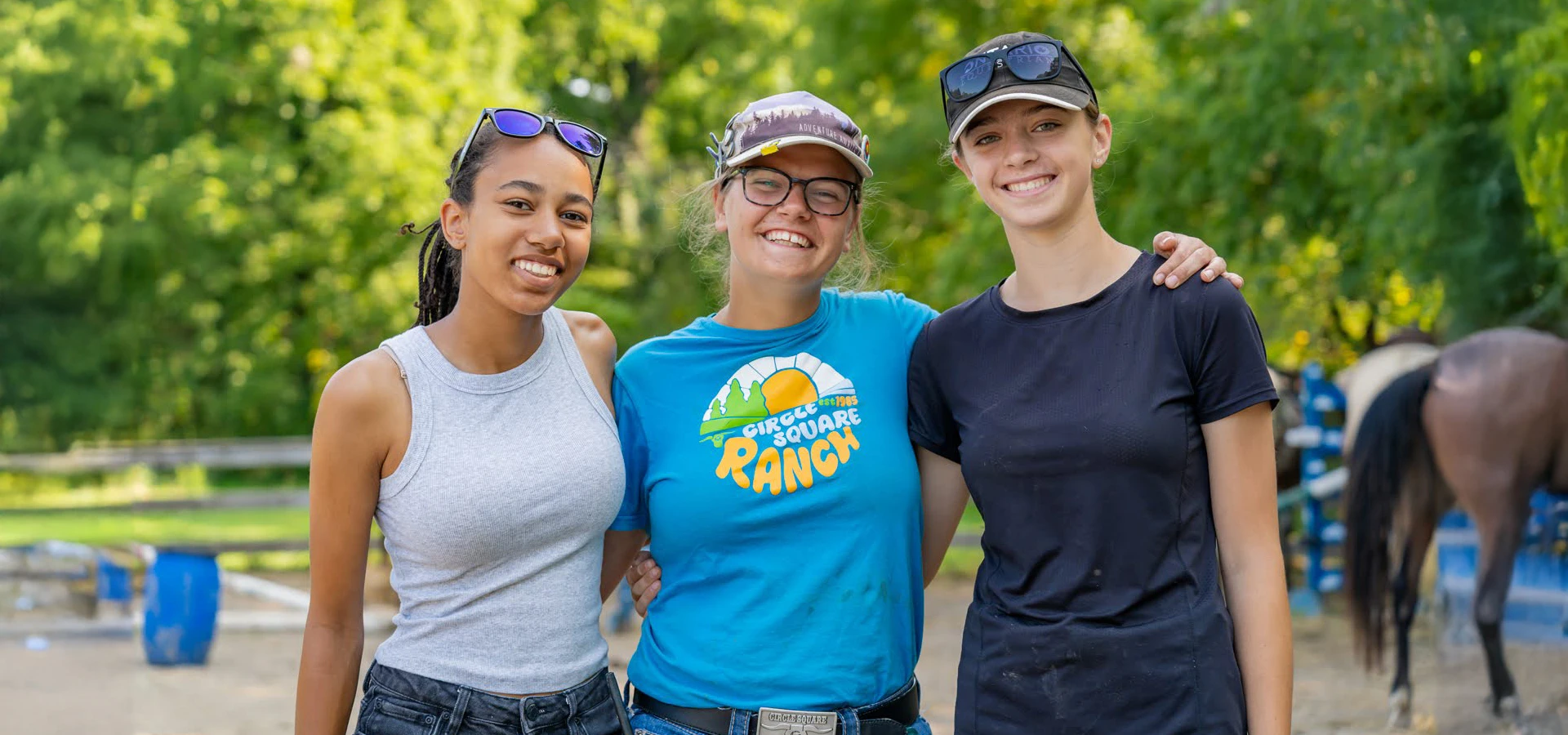 Three young women smiling and posing together outdoors at an equestrian ranch with a horse in the background.