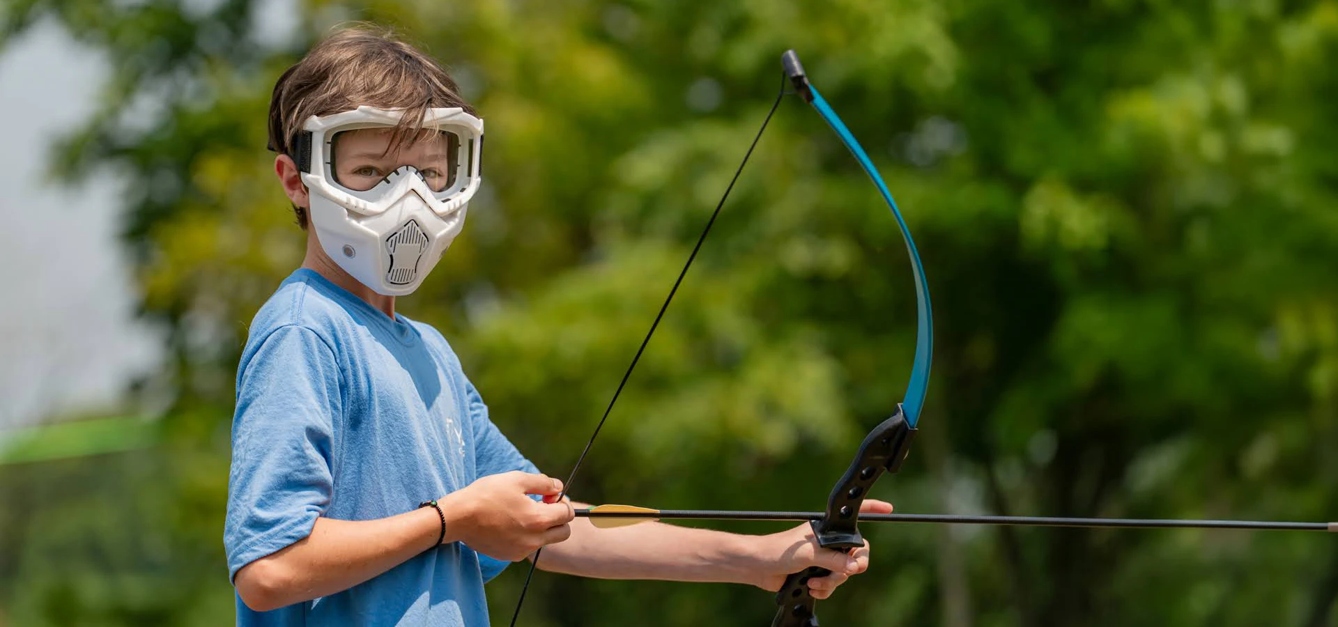 Young boy wearing protective goggles practices archery outdoors with a blue and black bow.