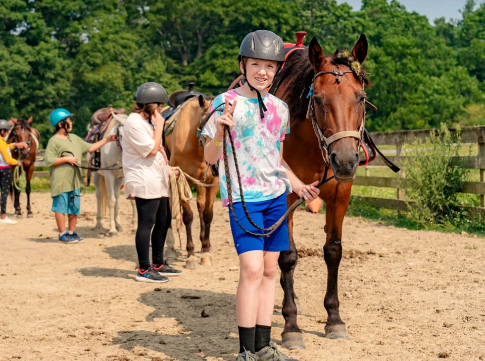 Young girl in a helmet leading a brown horse at a sunny outdoor horseback riding lesson with other children and instructors in the background.