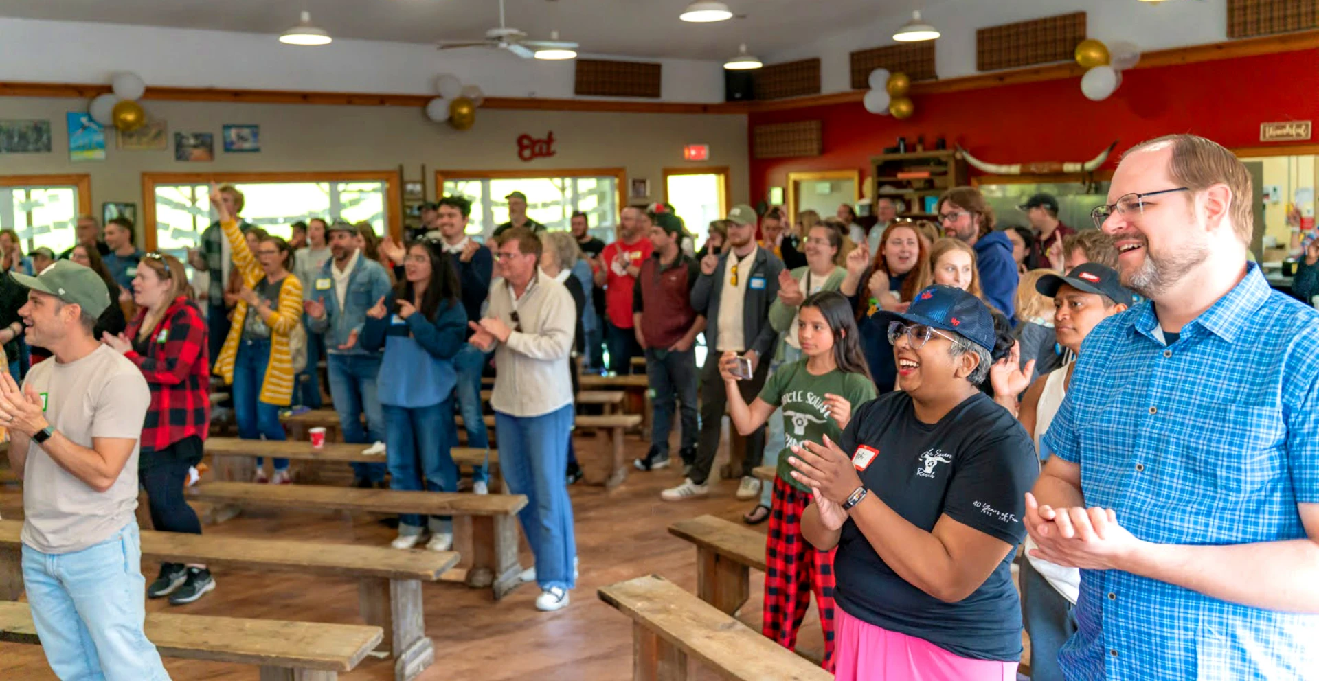 A diverse group of people standing and clapping inside a bright, spacious community hall with wooden benches.