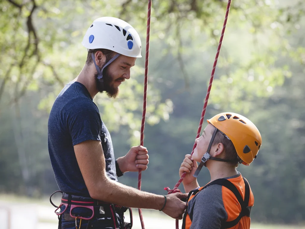 Man and boy wearing helmets and harnesses preparing for rock climbing outdoors with trees in the background.