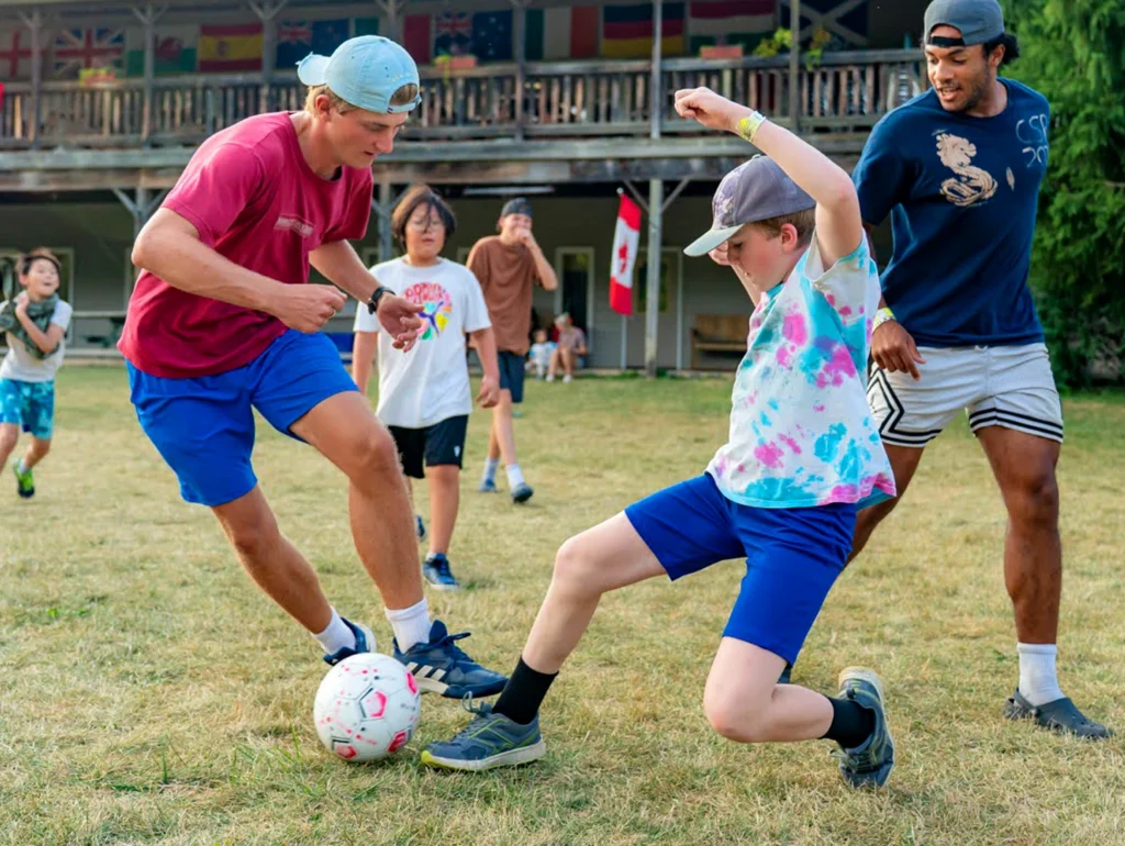 Children and a camp counselor playing soccer on a grassy field in front of a wooden lodge.