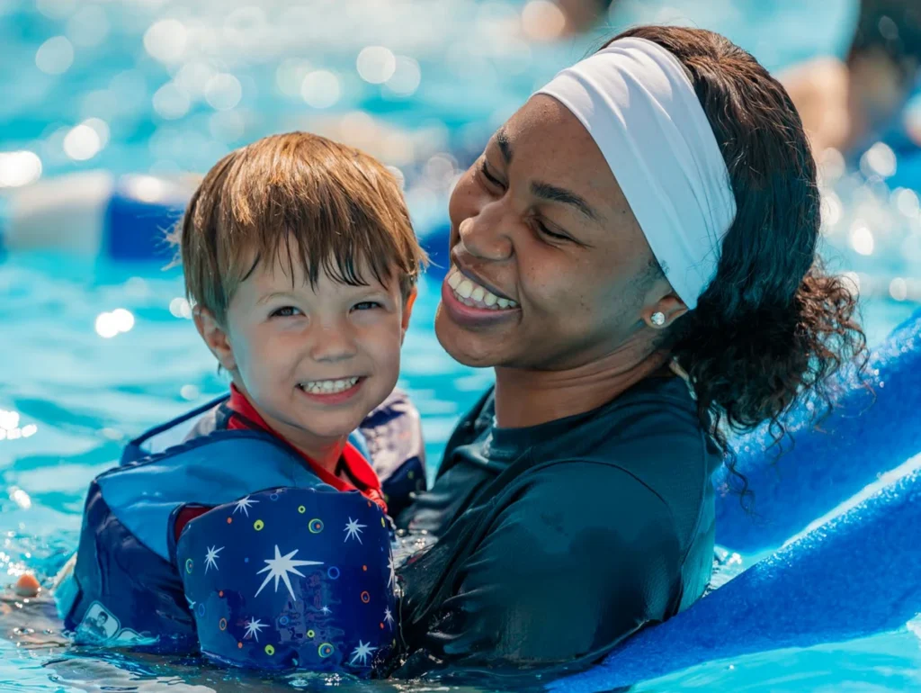 A smiling woman teaching a young child wearing a life jacket to swim in a pool.