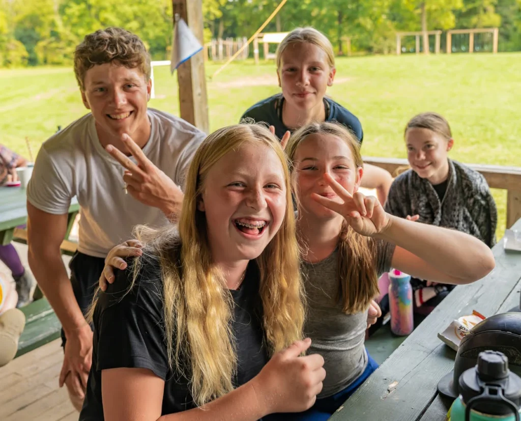 A group of five smiling teenagers sitting at an outdoor picnic table, enjoying a sunny day at camp.