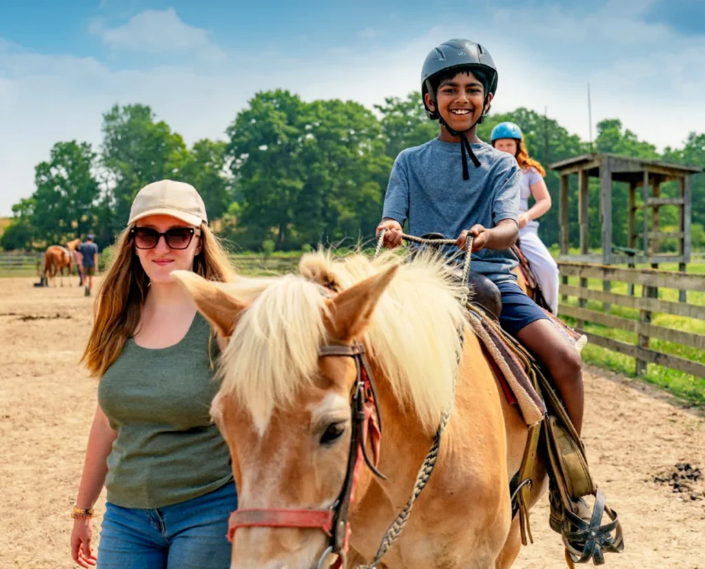 Smiling child wearing a helmet rides a light brown horse while an adult woman walks beside them on a sunny day at a horse stable.