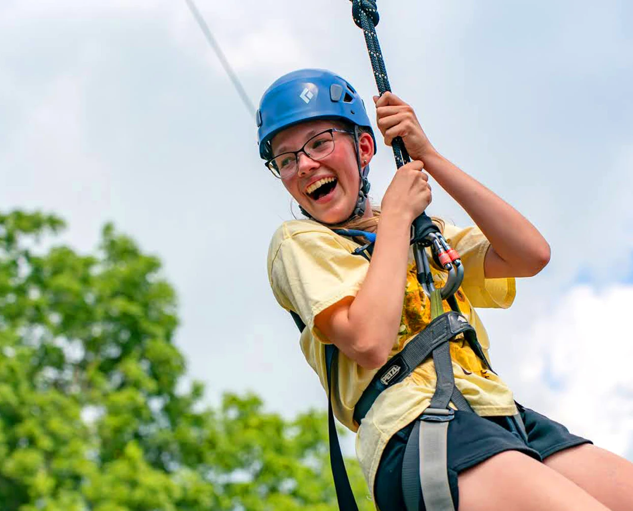 A teenager wearing a blue helmet and safety harness smiling while ziplining outdoors with green trees in the background.