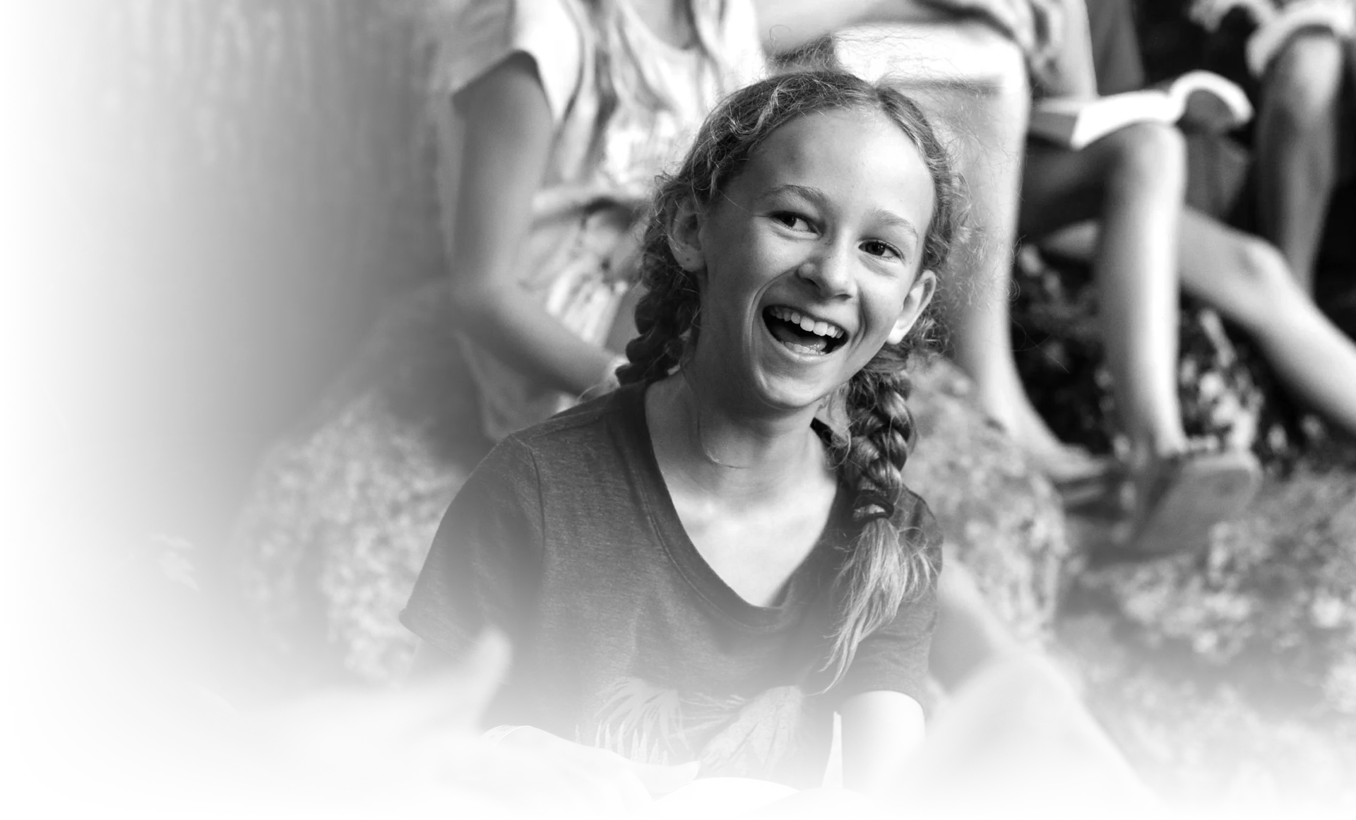 Black and white photo of a smiling young girl with braided hair sitting outdoors with other children.