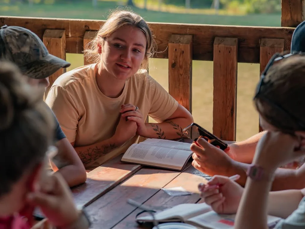 Group of young people engaged in a discussion around a wooden table outdoors, with notebooks and smartphones.