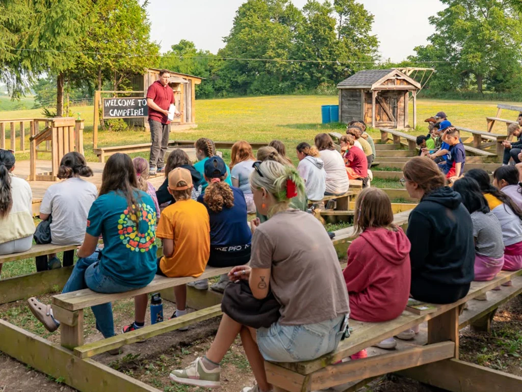A group of children seated on wooden benches outdoors attentively watching a person giving a presentation near a small wooden structure.