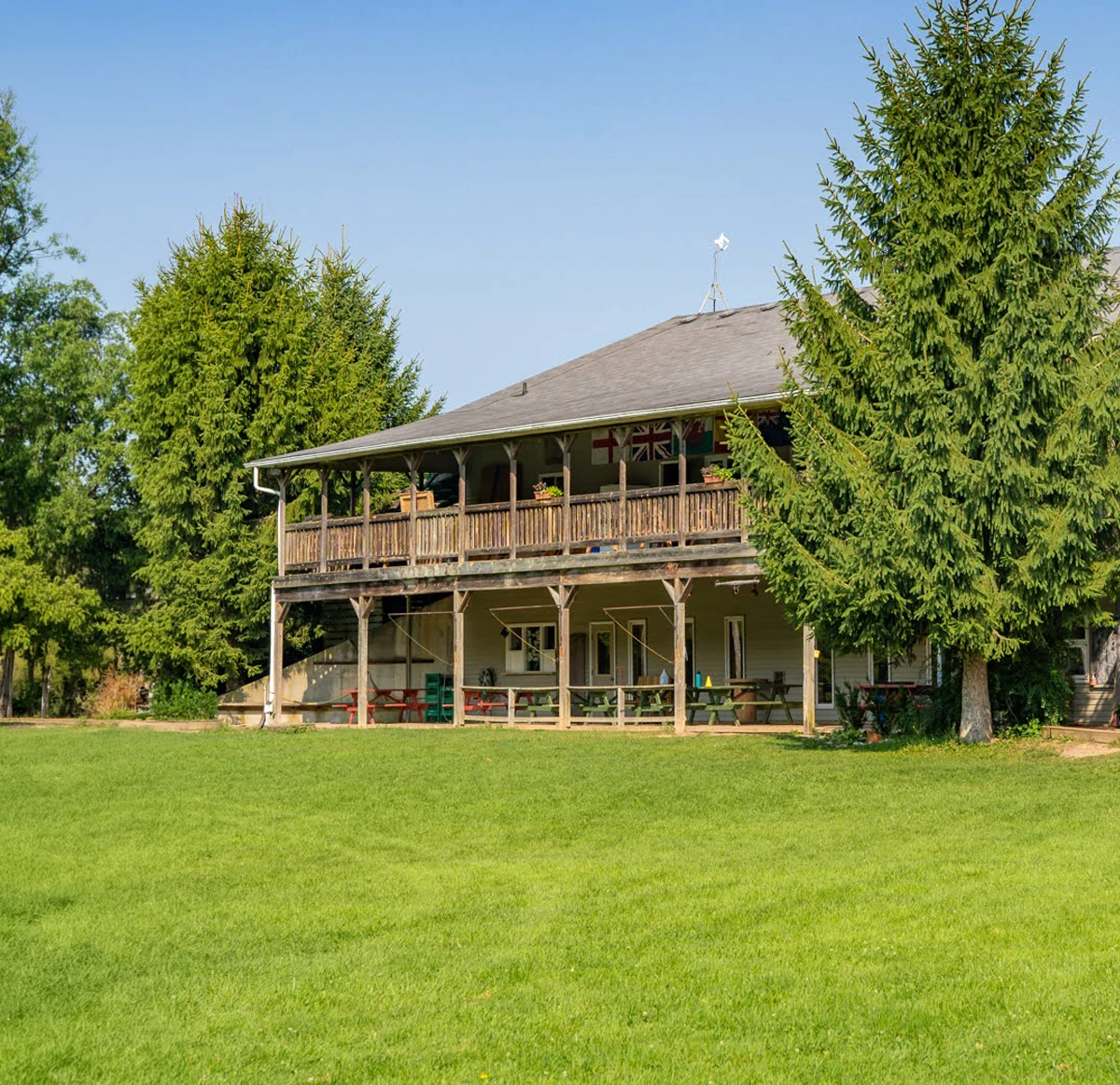Two-story wooden house with a large wraparound porch surrounded by tall evergreen trees and a green lawn under a clear blue sky.