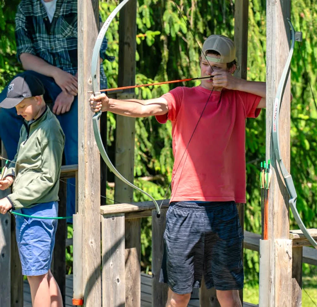 A young man in a red shirt and cap aims a bow and arrow at an outdoor archery range surrounded by green trees.