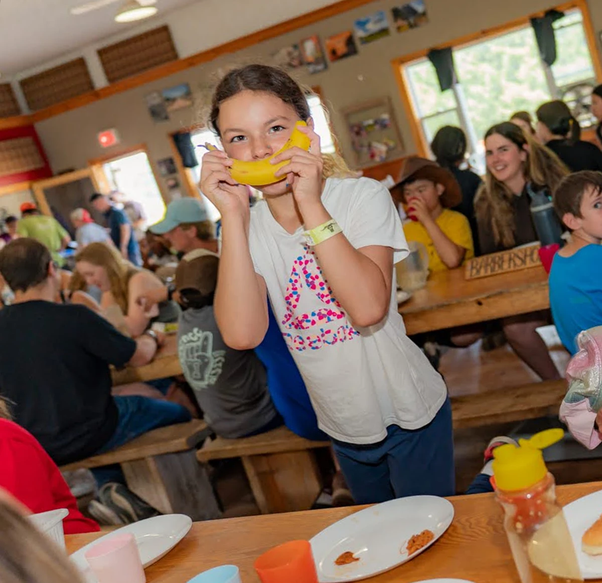 A young girl playfully holds a banana like a smile while sitting at a crowded cafeteria table with other children eating and socializing.
